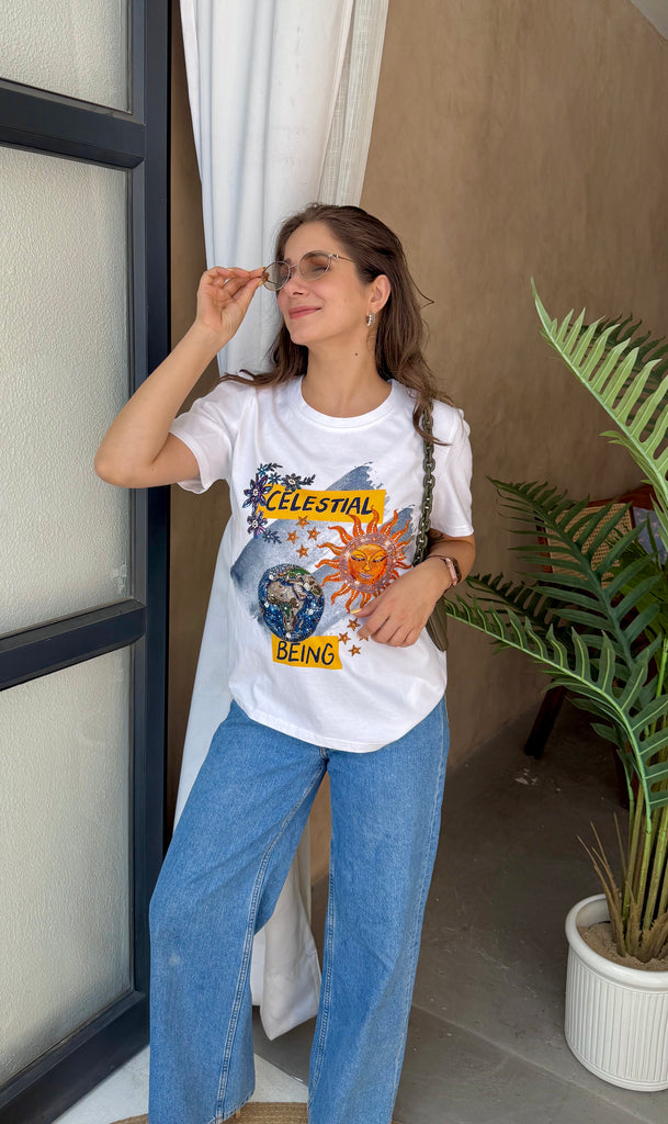 Woman wearing a white t-shirt with graphic design and text, standing indoors next to a plant.